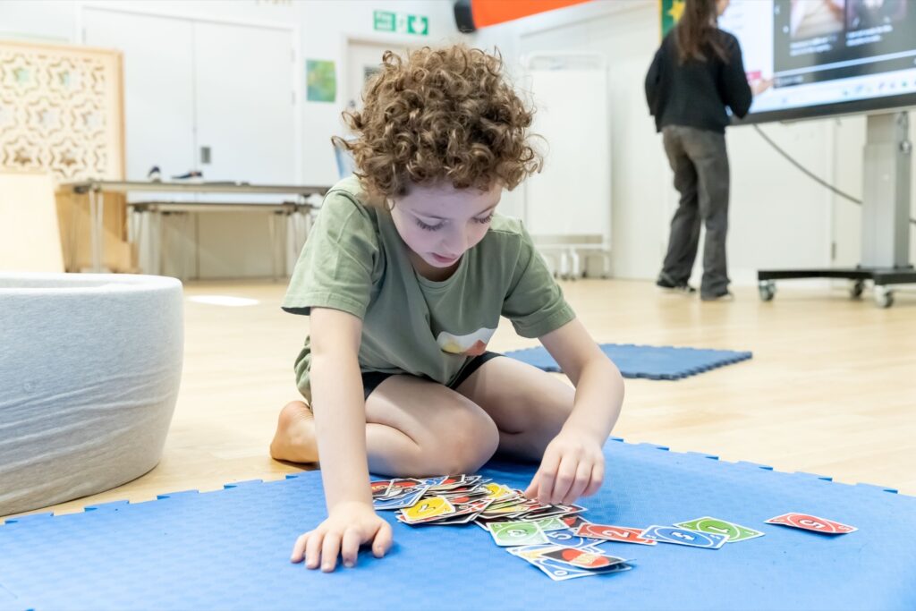 Child sitting on a blue mat, sorting UNO cards during an after school club activity while an instructor uses an interactive screen in the background.
